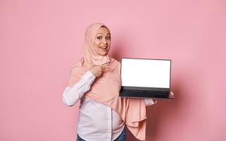 zwanger moslim vrouw in roze hijaab, points vinger Bij kopiëren ruimte voor advertenties Aan laptops wit leeg scherm, roze achtergrond foto