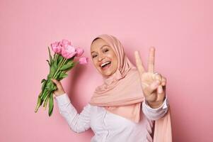 gelukkig moslim vrouw in hijaab, Holding boeket van tulpen, gebaren met handen, tonen vrede teken tegen roze achtergrond foto