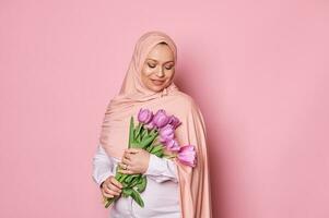 zelfverzekerd zwanger moslim vrouw in roze hijaab, glimlacht poseren met een boeket van Purper tulpen Aan geïsoleerd roze achtergrond foto