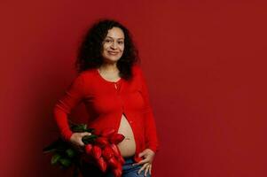 multi etnisch zwanger vrouw glimlacht op zoek Bij camera, houdt een boeket van rood tulpen Aan geïsoleerd kleur achtergrond foto