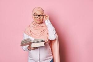 zelfverzekerd Arabisch moslim vrouw opvoeder charmant zwanger vrouw in roze hijab en bril, Holding boek, op zoek Bij camera foto