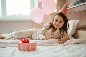 mooi kind meisje in feestelijk verjaardag hoed en pyjama, aan het liegen Aan de bed in haar slaapkamer en glimlachen op zoek Bij camera foto