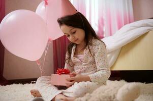 weinig verjaardag meisje in pyjama en roze feestelijk hoed, Holding schattig geschenk doos terwijl zittend Aan de tapijt in haar slaapkamer foto