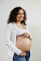 multi etnisch gekruld gravid vrouw Holding handen Aan haar buik, glimlachen schattig op zoek weg, geïsoleerd over- wit achtergrond foto