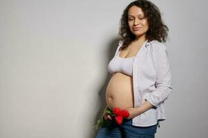 multi etnisch mooi zwanger vrouw ervan uitgaand een baby, lachend, poseren met rood tulpen Aan naakt buik, wit achtergrond foto