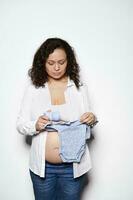 zwanger vrouw in tweede semester van zwangerschap, Holding blauw baby bodysuit, voorbereidingen treffen naar kind geboorte, geïsoleerd Aan wit foto