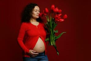 mooi spaans vrouw met een boeket van rood tulpen, aanraken haar buik poseren met haar ogen Gesloten Aan rood achtergrond foto