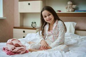 gelukkig weinig kind meisje in pyjama- zittend Aan de bed in de buurt haar gevouwen kleren glimlachen schattig op zoek Bij camera foto
