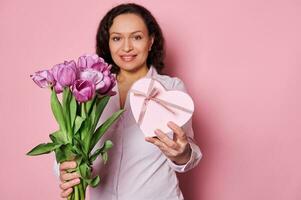 details Aan boeket van Purper tulpen en hart vormig geschenk doos in handen van wazig gelukkig glimlachen vrouw, geïsoleerd Aan roze foto