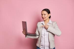 charmant Afrikaanse Amerikaans bedrijf vrouw manager points Bij laptop computer, glimlacht op zoek Bij camera, geïsoleerd Aan roze foto