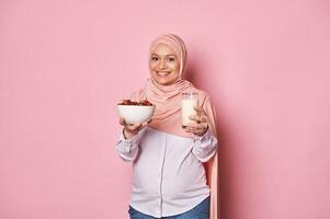 moslim zwanger vrouw in hoofd sjaal, Holding glas van gezond veganistisch melk en kom van datums, ideaal ontbijt in Ramadan foto