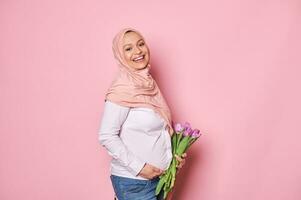 gelukkig moslim zwanger vrouw in roze hijaab, glimlachen met mooi glimlach, aanraken haar buik, geïsoleerd Aan roze achtergrond foto