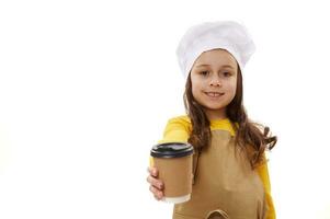 lief weinig kind meisje in chef-kok hoed en schort, Holding uit Bij camera een meenemen heet drinken in beschikbaar karton kop foto