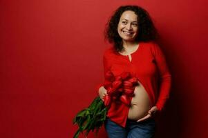 verrukkelijk etnisch zwanger vrouw houdt rood tulpen, aanraken haar buik, mysterieus glimlacht op zoek terzijde, rood achtergrond foto