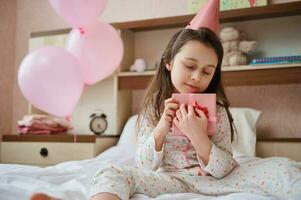 aanbiddelijk baby meisje in pyjama en roze feestelijk hoed, voorzichtig knuffels haar verjaardag Cadeau, zittend met Gesloten ogen Aan bed foto