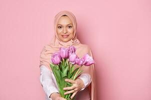 mooi Arabisch moslim vrouw in roze hijaab, Holding uit Bij camera een schattig boeket van tulpen, glimlachen op zoek Bij camera foto