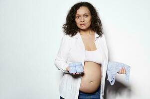 portret van zwanger vrouw Aan tweede trimester van zwangerschap, poseren met blauw pasgeboren bodysuit en baby schoenen Aan wit foto