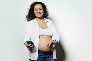 glimlachen zwanger vrouw records weeën of baby bewegingen Aan mobiel app in haar smartphone, glimlachen breed Bij camera foto