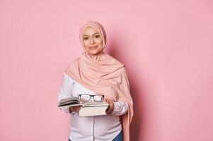 zelfverzekerd moslim vrouw opvoeder in roze hijaab, een school- leraar Holding bril en aan het studeren boek, roze achtergrond foto