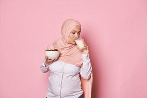 lief moslim zwanger vrouw in roze hijaab, Holding een boog met datums en drinken gezond veganistisch melk, geïsoleerd Aan roze foto