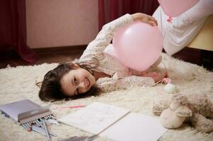 weinig meisje in pyjama, aan het liegen Aan een tapijt in haar slaapkamer, spelen met roze pastel ballonnen, glimlachen op zoek Bij camera foto