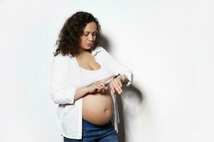 geïsoleerd portret Aan wit achtergrond van gekruld haren verwachtend vrouw tellen weeën Aan haar slim pols kijk maar foto