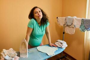 uitgeput zwanger vrouw in 3d trimester van zwangerschap, staand door strijken bord, stomen pasgeboren baby kleren foto