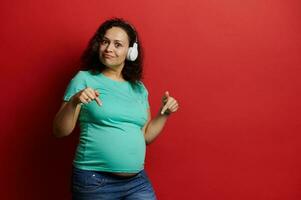 charmant volwassen zwanger vrouw tonen haar buik naar camera, luisteren naar muziek- Aan draadloze hoofdtelefoons over- rood achtergrond foto