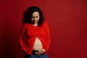 Afrikaanse Amerikaans positief zwanger vrouw aanraken haar buik, lachend, geïsoleerd Aan rood achtergrond met kopiëren advertentie ruimte foto