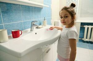 aanbiddelijk weinig meisje Holding tandenborstel, looks Bij camera staand door wastafel in de badkamer gedurende ochtend- hygiëne routine- foto