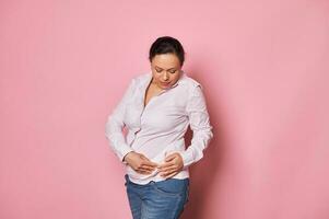 zelfverzekerd zwanger vrouw zetten Aan orthopedische buik ondersteunen verband, geïsoleerd over- roze achtergrond foto