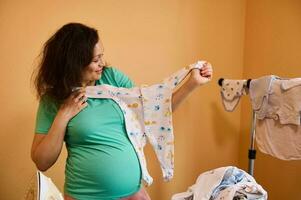 multi etnisch zwanger vrouw in huis draagt, Holding een klein schattig baby kleding, voorbereidingen treffen uitrusting voor pasgeboren bevalling foto