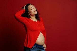 mode portret van multi etnisch mooi zwanger vrouw, poseren met Gesloten ogen en kaal buik over- rood achtergrond foto