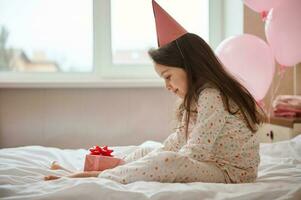 weinig meisje in pyjama en feestelijk roze hoed, zittend Aan de bed in haar slaapkamer en Holding haar verjaardag Cadeau in handen foto