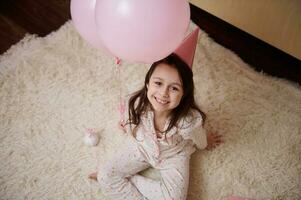 top visie lief weinig kind, verjaardag meisje in pyjama en roze partij hoed, zittend Aan tapijt, glimlachen op zoek Bij camera foto
