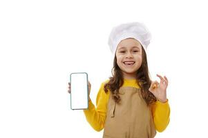 positief weinig kind meisje in chef hoed en schort, tonen smartphone met blanco wit scherm. kopiëren ruimte voor mobiel apps foto