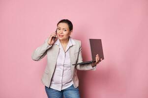 zwanger bedrijf vrouw, Holding laptop, pratend Aan mobiel telefoon, uitdrukken teleurstelling, geïsoleerd roze achtergrond foto