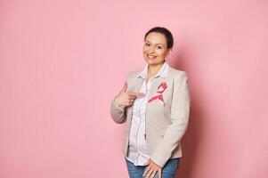 positief multi-etnisch vrouw, staand over- roze achtergrond en richten haar vinger Bij een roze satijn lint Aan haar jasje foto