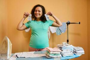 zwanger vrouw houdt pasgeboren sokken, glimlacht Bij camera staand Bij strijken bord met gestreken baby kleren foto