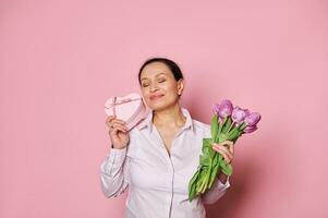 portret van een verrukkelijk vrouw, moeder gevoel aangeraakt krijgen gelukkig Cadeau en boeket van tulpen voor speciaal gelegenheid foto