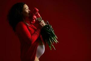 kant portret Aan rood achtergrond van mooi zwanger vrouw snuiven een boeket van rood tulpen. gelukkig moeder dag concept foto