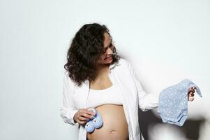 volwassen zwanger vrouw houdt blauw bodysuit en gebreid baby slofjes voor toekomst pasgeboren, voorbereidingen treffen dingen in zwangerschap tijd foto