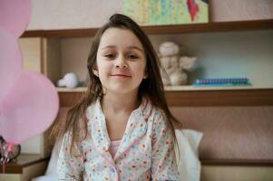 Kaukasisch weinig 5 jaren oud kind meisje in pyjama, glimlachen op zoek Bij camera, zittend Aan de bed in haar aantrekkingskracht slaapkamer foto