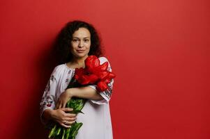 mooi vrouw in geborduurd wit jurk, houdt een bundel van rood tulpen, glimlacht op zoek Bij camera, geïsoleerd rood backdrop foto