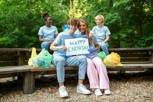 gelukkig vrijwilligers Holding aanplakbiljet met 'Vrolijk aarde dag' bericht. vrijwilligerswerk, liefdadigheid, schoonmaak, mensen en ecologie concept - groep van gelukkig vrijwilligers met vuilnis Tassen schoonmaak Oppervlakte in park foto