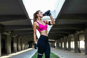 mooi vrouw loper staand buitenshuis drinken water van haar fles. geschiktheid vrouw nemen een breken na rennen training.atletisch jong vrouw in sport- jurk aan het doen geschiktheid oefening. foto