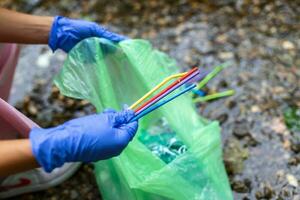 hand- van vrouw plukken omhoog plastic rietjes in vuilnis Tassen terwijl schoonmaak klein rivier. vrijwilligerswerk, liefdadigheid, mensen, ecologie concept. detailopname vrijwilliger verzamelen plastic uitschot in Woud. foto