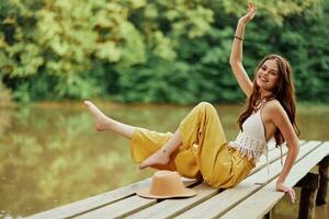 een jong hippie vrouw zit Aan een meer brug vervelend elegant eco kleren en glimlachen foto