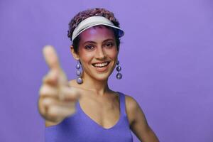 portret van een sportief mode vrouw poseren glimlachen met tanden en richten een vinger Bij de camera in een Purper yoga trainingspak en een transparant pet Aan een Purper monochroom achtergrond foto