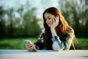 een vrouw met gekruld rood haar- in een plaid overhemd zit buitenshuis in de park met een cel telefoon in haar handen en looks weg. freelance werk in de park foto